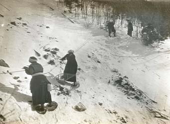 Women in long dresses and snowshoes climb a steep slope.