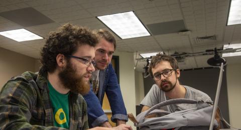 Two students look intently at a laptop screen with a professor.