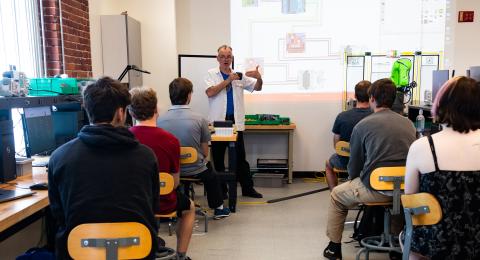 A professor stands in the front of a classroom with six students watching. The projection behind the professor displays a detailed electronic schematic.