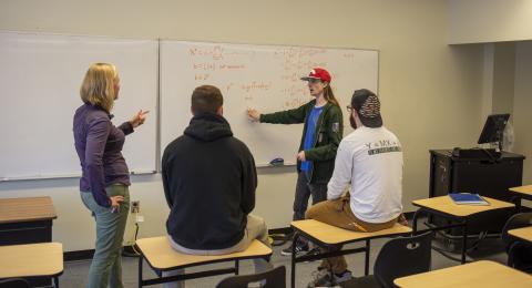 A mathematics professor with 3 students works on an equation written on dry erase board.