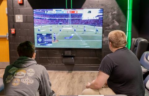 Two male students sit in the eSports lounge playing a video game together.