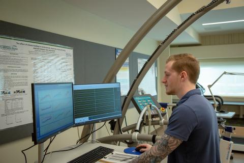A white man sits at a desk in the Human Performance Center's Research lab. He is looking at two computer monitors and holds a computer mouse in his hand.