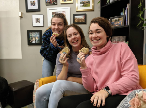Three students sit in the Office of Community Impact smiling while they hold cookies.