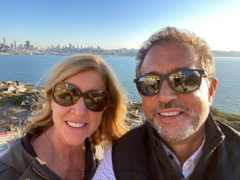 Photo of a man, David Almeda, and his wife, Erin wearing sunglasses and water and city skyline standing in front of a view of the