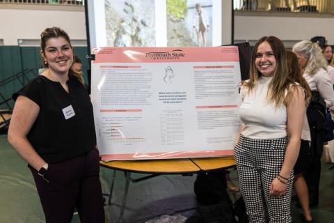 Two female students standing beside a large display board featuring their showcase project.