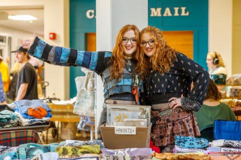 Two women pose for a photo in front of a bin sitting on a table labeled "Prints". They are joined together to celebrate PSU's Earth Jam event.