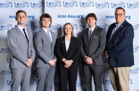 Logo for the website ncsc-ksu.org. Four men stand on either side of a woman in business attire, posing for a photo.