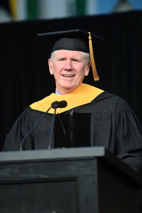 A man wearing a graduation cap and gown stands at a podium and speaks into a microphone.