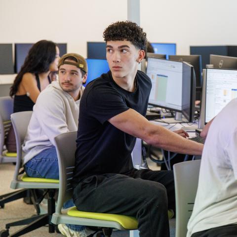 A student sits in front of a computer, turned towards an individual who is speaking.