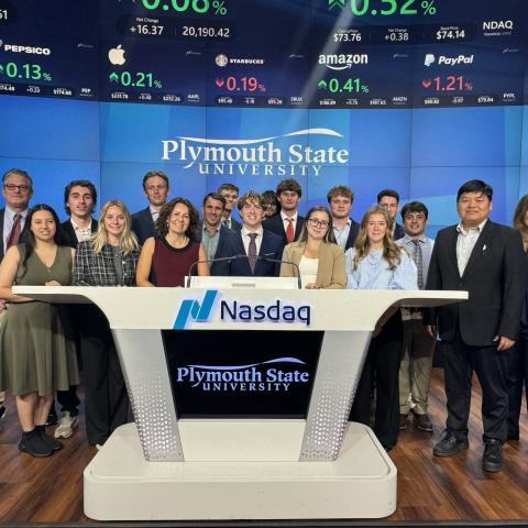 A group of indiduals post for a photo under a ticker. The podium shows a "NASDAQ" logo, along with a Plymouth State University logo.
