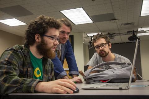 Two students look intently at a laptop screen with a professor.