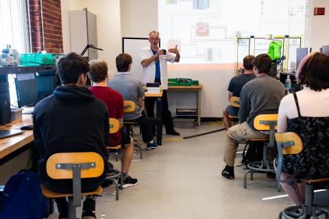 A professor stands in the front of a classroom with six students watching. The projection behind the professor displays a detailed electronic schematic.