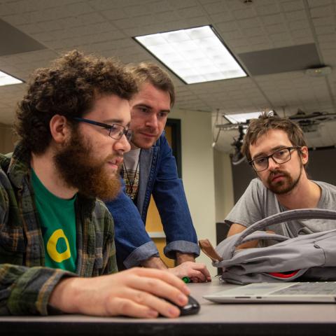 Two students look intently at a laptop screen with a professor.